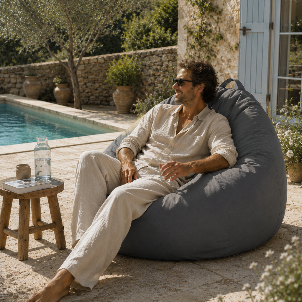 Homme assis sur un pouf extérieur gris Mowana au bord d’une piscine avec ambiance terrasse méditerranéenne premium